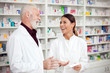 © Ivan - Medicine, pharmaceutics, health care and people concept - Young female and senior male pharmacists smiling and standing in front of shelves with medications