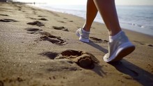Close-up Of Woman Foot In Sneakers Walking Along The Sea On A Sandy Beach Early In The Morning On Vacation