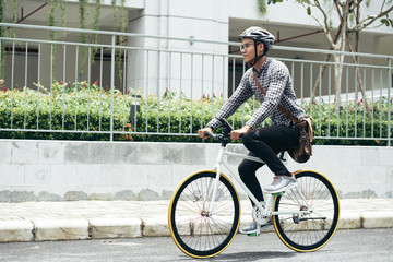 Young Asian man wearing casual clothes, glasses and safety helmet riding bicycle along the road while going home after university