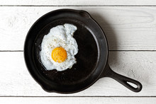 Single Fried Egg In Cast Iron Frying Pan Sprinkled With Ground Black Pepper. Isolated On White Painted Wood From Above.
