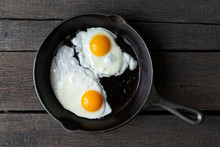 Two Fried Eggs In Cast Iron Frying Pan Isolated On Dark Painted Wood From Above.
