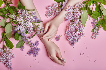 Wall Mural - Fashion art hands natural cosmetics women, bright purple lilac flowers in hand with bright contrast makeup, hand care. Creative beauty photo of a girl sitting at table on contrasting pink background