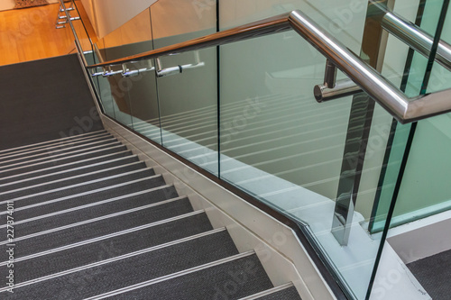 Stairs are leading downward to a small platform. The stair steps are gray. A metal handrail and glass panels are next to the stairs.