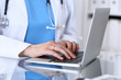© rogerphoto - Doctor typing on laptop computer while sitting at the glass desk in hospital office. Physician at work. Medicine and healthcare concept