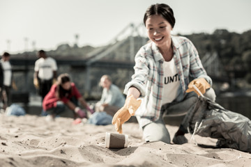 Gathering cups. Smiling socially active woman wearing bright yellow gloves gather empty coffee cups on beach