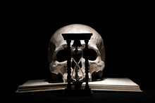 Human Skull On Old Open Book With Vintage Hourglass On Black Background. Drama And Time Concept.