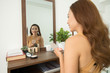 © DragonImages - miling beautiful Vietnamese lady sitting in front of mirror and applying makeup while getting ready for work