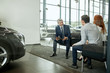 © alfa27 - Sales agent at the table desk telling about car features to cheerful couple of customers at car dealership office