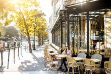 Street View On The Traditional French Cafe With Young Woman Sitting Outdoors During The Morning Light In Paris
