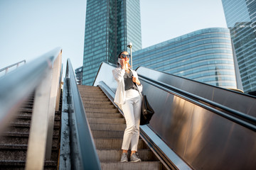 Wall Mural - Stylish businesswoman in white suit talking phone while going down on the escalator at the business centre outdoors in Paris