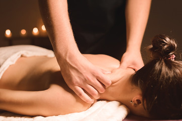 Wall Mural - Men's hands make a therapeutic neck massage for a girl lying on a massage couch in a massage spa with dark lighting. Close-up. Dark Key