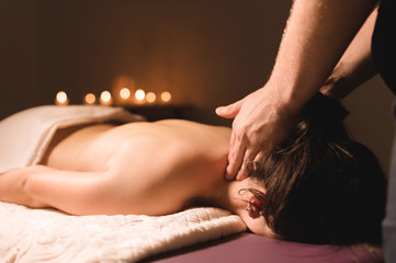 Men's hands make a therapeutic neck massage for a girl lying on a massage couch in a massage spa with dark lighting. Close-up. Dark Key