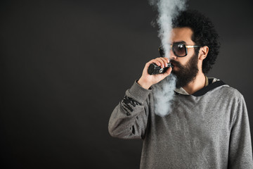 Wall Mural - Vaping man holding a mod. A cloud of vapor. Black background. Vaping an electronic cigarette with a lot of smoke. Vape concept