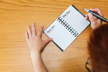 Woman fills in her goals and plans for long and short terms in spiral notepad on wooden table, top view