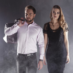 Young handsome man and pretty woman walking in smoke. Black dress, white shirt, leather hand bag. Dark studio background