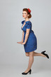Vertical picture of gorgeous redhaired young woman wearing black high heeled shoes and dotted blue dress with low neck cut posing in studio, holding hands in front of her and keeping leg raised