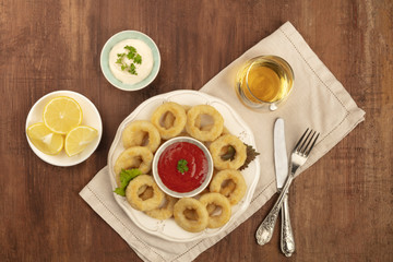 Squid rings with two sauces, lemon wedges, white wine, and copy space, shot from the top on a dark wooden background with vintage cutlery