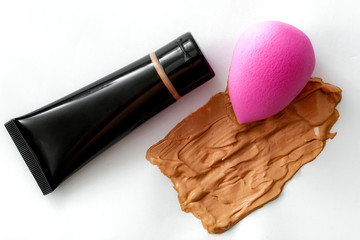 Face foundation application tool and makeup products concept with a pink beauty blender sponge, plastic tube of liquid foundation and smeared face product isolated on a white background