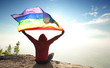 © 10incheslab - man sitting on mountain top raised rainbow LGBT symbol flag to bright sunny blue sky