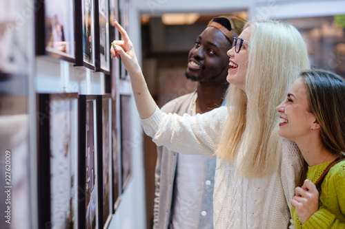 Fotografía Three young people hipster students with different skins looking happy together, talkng, looking at picture on wall, spending free time together in art photo gallery