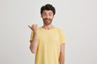 © timtimphoto - Portrait of smiling glad attractive young man with dark brown hair and beard wears casual yellow tshirt, points left with one thumb, standing center isolated over white background