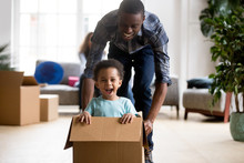 Black Family In Living Room Have A Fun Spend Time At New Home. African Adorable Playful Laughing Boy Sitting At Cardboard Box, Father Rolling Him Playing Together. New Property And Relocation Concept