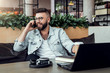 © foxyburrow - Bearded businessman,blogger sitting in cafe,talking on smart phone,working on laptop, freelancer working in coffee shop.