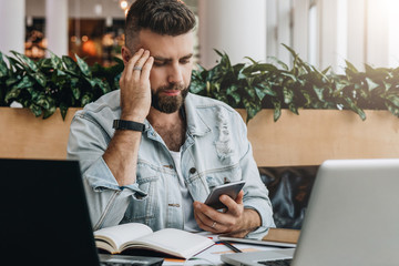 Hipster man uses smartphone, works on two laptops. Businessman concentrates reads an information message in phone.