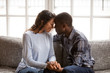 Black african married couple in love together at home. American woman and man embrace enjoy company of each other sitting on couch holding hands having romantic date. Feelings and first kiss concept