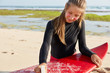 Cropped shot of attractive woman uses wax for safe surfing, has appealing appearance, dressed in wetsuit, poses on beach near bay, recreats in tropical country. Extreme sport and lifestyle concept