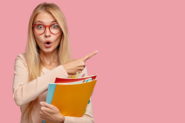 Photo of surprised teacher indicates aside with index finger, has light straight hair, keeps mouth opened with amazement, carries textbooks, isolated on pink background. Omg, look at this space