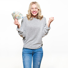 Super Excited Young Woman In Grey Sweater And Jeans Holding Bunch Of Euro Banknotes, Clinching Fists, Celebrating Winning Lottery. Ecstatic Woman Holding Lots Of Money, Isolated On White Background.