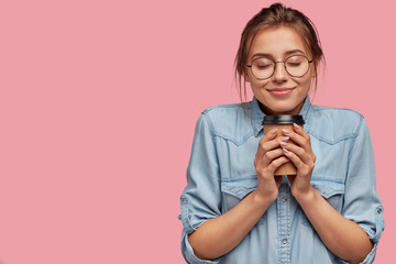 Wall Mural - Pleased lovely Caucasian woman holds aromatic beverage, drinks cappuccino or coffee, feels warm, closes eyes from pleasure, has charming smile, wears denim shirt, isolated over pink background
