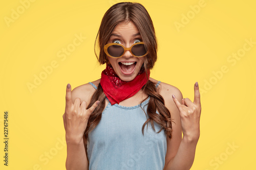Foto Headshot of provocative cool female hipster shows rock n roll hand gesture, exclaims something loudly, wears fashionable bandana, trendy sunglasses, poses against yellow background