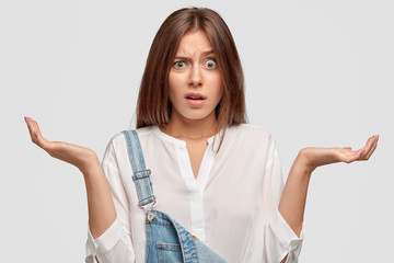 Canvas Print - Photo of indignant European woman raises eyebrows, shrugs shoulders, dressed in white shirt and denim overalls, has clueless expression, models over white background, asks why she should prepare task