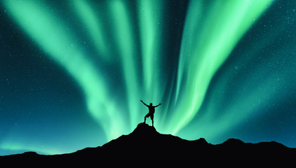 Wall Mural - Northern lights and silhouette of standing man with raised up arms on the mountain in Norway. Aurora borealis and happy man. Sky with stars and green polar lights. Night landscape with aurora. Concept