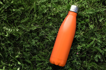 Stainless thermos water bottle isolated on green grass outdoor. Orange matte color. Horizontal photo without effects.