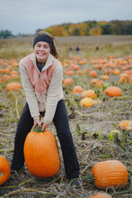 Adult Woman (30s) Attempts To Pick Up A Giant Pumpkin From A Pumpkin Patch. Smiling And Laughing And Having Fun
