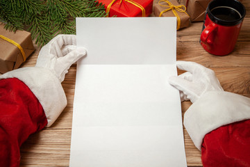 Santa Claus holding letter on wooden table with gift boxes and Christmas tree and cup of hot coffee or tea.  mockup blank