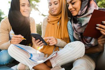 Canvas Print - Photo of attractive muslim girls wearing headscarfs, sitting on blanket in green park and studying with laptop and smartphone