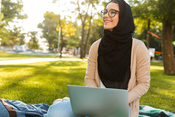 Wall Mural - Photo of happy arabic woman wearing headscarf, sitting on blanket in green park and using silver laptop