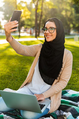 Wall Mural - Photo of pretty arabic woman wearing headscarf using silver laptop, and taking selfie in green park