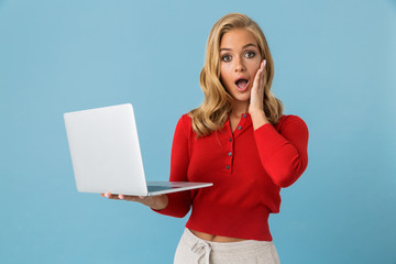 Poster - Portrait of shocked or excited woman 20s holding silver laptop, isolated over blue background in studio