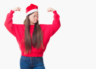 Wall Mural - Young Chinese woman over isolated background wearing christmas hat showing arms muscles smiling proud. Fitness concept.