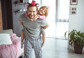 Happy family. Portrait of excited girl sitting on daddy back and laughing. Man is standing at home and looking at camera with cheerfulness. He wear makeup and rollers on hair 