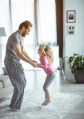 Wall Mural - Lets play. Excited little girl is jumping while holding hands of her father. She is laughing. Home entertainment concept 