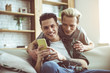 © Yakobchuk Olena - Toned portrait of dark-haired guy in headphones sitting on couch and holding cellphone while his boyfriend snuggling up to him from behind
