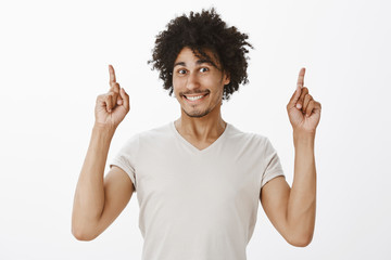 Wall Mural - Studio shot of charming mature hispanic male hipster with curly hair and moustache smiling joyfully while gazing at camera with excitement, pointing up with index fingers while posing over grey wall