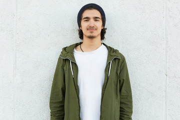 Horizontal shot of stylish teenage boy wears black hat, white t shirt and green anorak, looks with satisfied expression direclty at camera, isolated over white background. Lifestyle and people concept