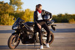 Photo of young female motorcyclist wears fashionable clothes, white sneakers, holds helmet under arm, stands near black motorbike, stands on asphalt, poses outdoor. People and lifestyle concept